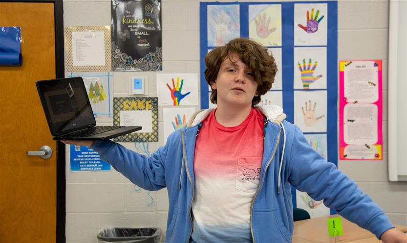 A student wearing a red, white and blue shirt holds a laptop in the air.